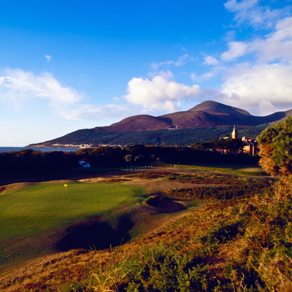 Golf course photograph of Royal County Down in Newcastle, Northern Ireland, showing the ninth hole with clubhouse, hotel, and dramatic clouds over the mountains.
